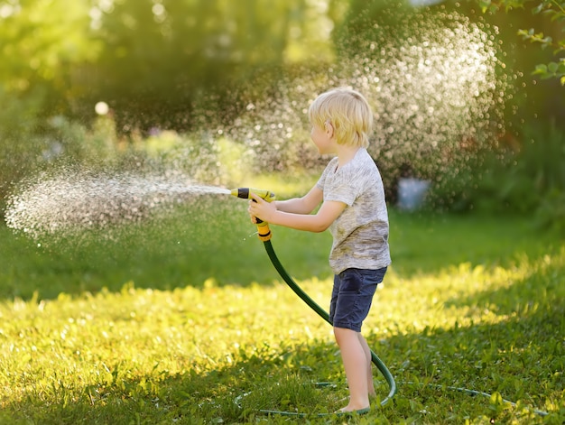 Rapaz pequeno engraçado que joga com a mangueira de jardim no quintal ensolarado. Criança de pré-escola se divertindo com spray de água.