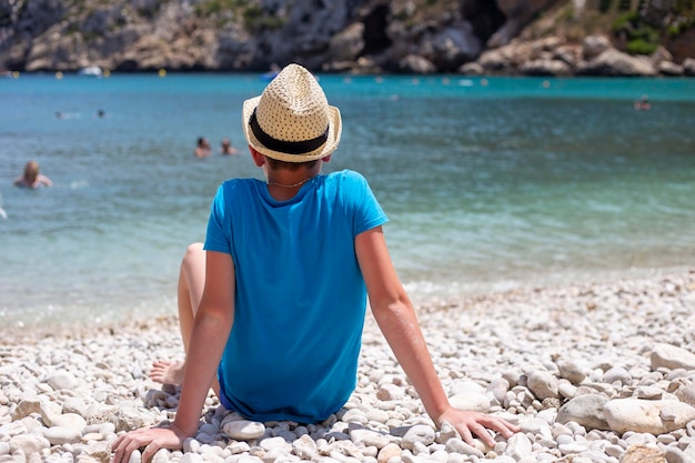 Rapaz sem rosto em t-shirt azul e chapéu de palha senta-se sozinho na praia tropical