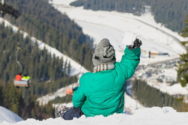 Rapaz senta-se na encosta nevada de uma colina e joga bola de neve