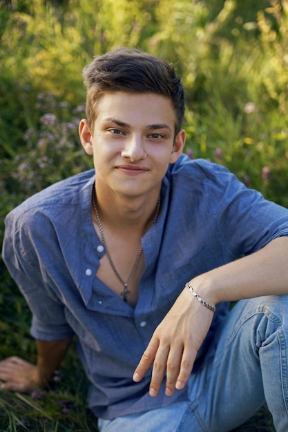 Rapaz sexy adolescente sentado na grama na natureza em uma camisa e calça jeans descalço e olhando para a câmera