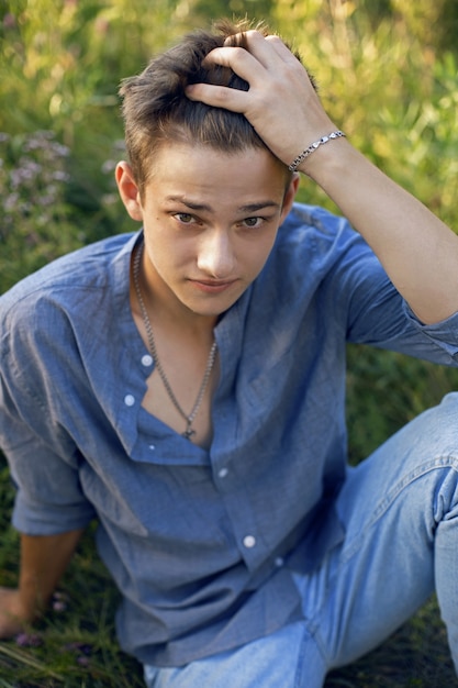 Rapaz sexy adolescente sentado na grama na natureza em uma camisa e calça jeans descalço e olhando para a câmera