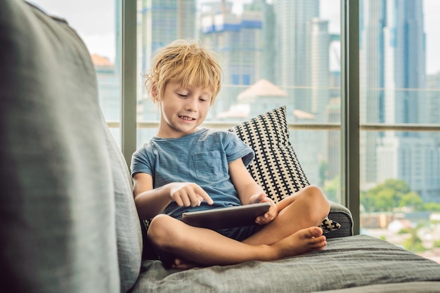 Rapaz usa um tablet em casa no sofá no fundo de uma janela com arranha-céus. Crianças modernas na megalópole usam um conceito de tablet