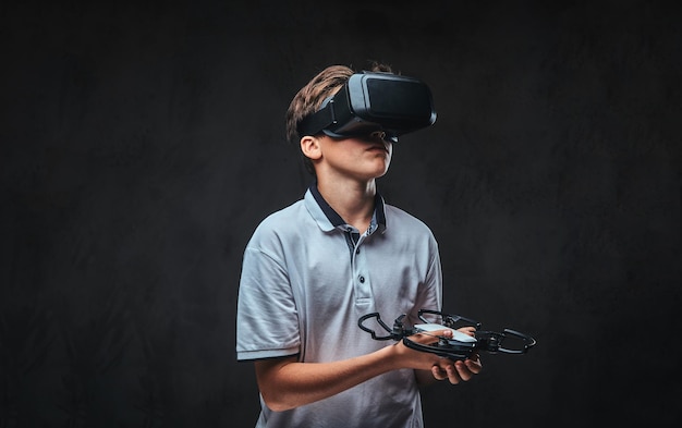 Rapaz vestido com uma camiseta branca usando óculos de realidade virtual e segura um quadricóptero. Isolado em um fundo escuro.