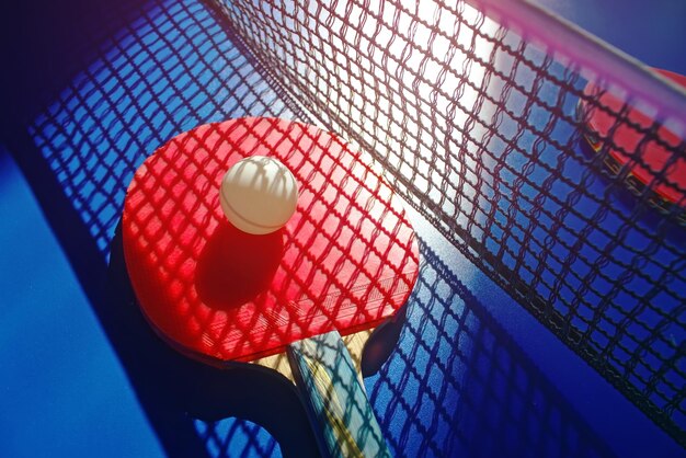 Una raqueta de tenis de mesa roja y una pelota blanca yacen en la superficie de la mesa junto a la red Juego deportivo