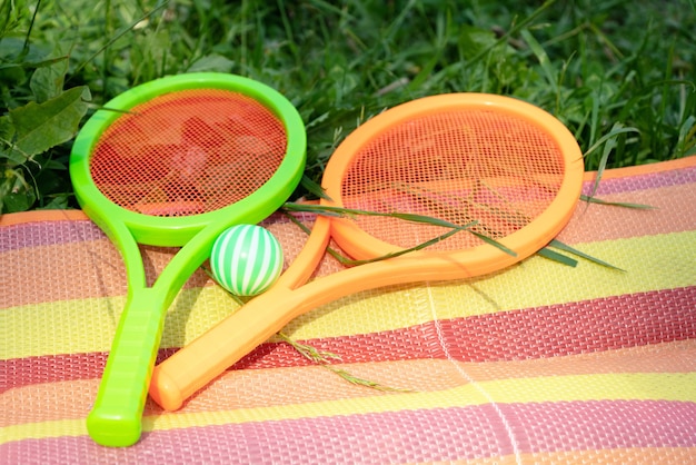 Foto raquetes de tênis de brinquedo com a bola deitada em uma manta listrada colorida espalhada na grama