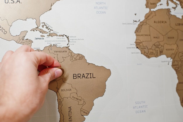 Rasca el mapa de viaje del mundo, la mano del hombre borra Brasil con monedas.