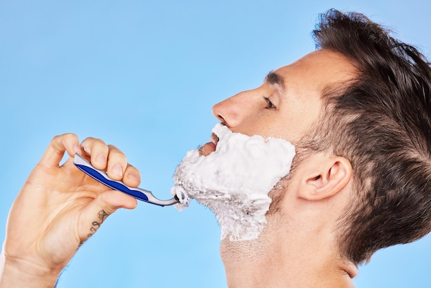 Rasiercreme und Mann, der seinen Bart mit einem Rasierer im Studio für Gesundheit, Hautpflege und Hygiene pflegt Schaumrasierer und Modell rasieren sein Gesichtshaar für eine Gesichts- oder Hautroutine, die durch blauen Hintergrund isoliert ist