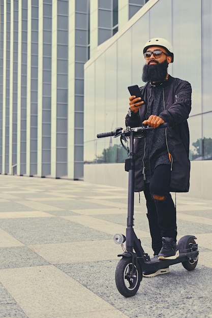 Rasierter Kopf tätowiert, bärtiger Mann mit einem Smartphone über modernem Gebäudehintergrund nach der Fahrt mit dem Elektroroller.