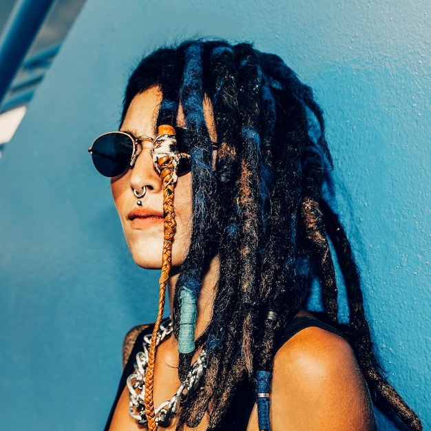 Rasta Spanish Girl com dreadlocks, piercings, tatuagens e acessórios estilosos na parede azul.