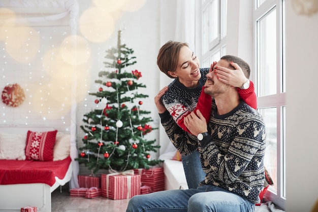 Rate mal, was ich dir schenken werde. Glückliche junge Leute sitzen auf der Fensterbank im Raum mit Weihnachtsdekorationen