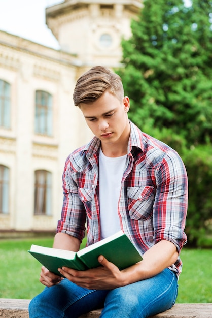 Ratón de biblioteca guapo. Libro de lectura de estudiante masculino seguro mientras está sentado en el banco y frente al edificio de la universidad