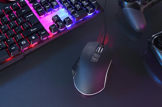 El ratón de la computadora, el teclado RGB y los controladores de juegos en luces de neón en fondo oscuro por encima de la vista