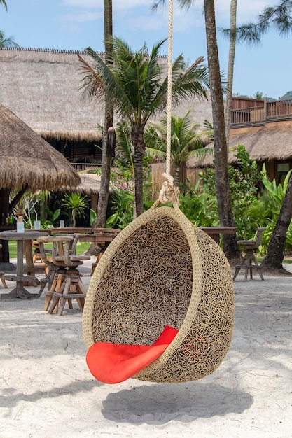 Rattan-Schaukelstuhl, der an einem Baum an einem tropischen Sandstrand in der Nähe des Meeres hängt Reise- und Urlaubskonzept Thailand
