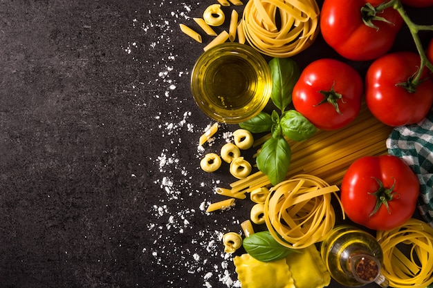 Ravioles, pasta penne, espagueti, tortellini, tomate y albahaca sobre negro. Vista superior, espacio de copia