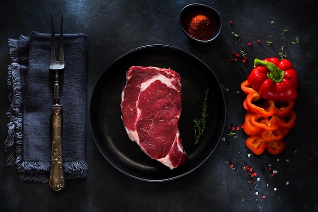 Raw Steak Ribeye em prato, garfo de carne, ervas e especiarias em um fundo escuro de concreto