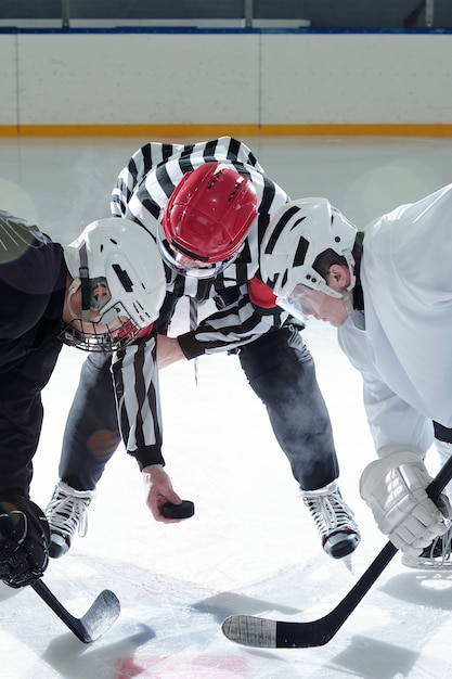 Árbitro de hockey sosteniendo el disco sobre la pista de hielo con dos rivales con palos de pie a su derecha e izquierda y esperando el momento para disparar