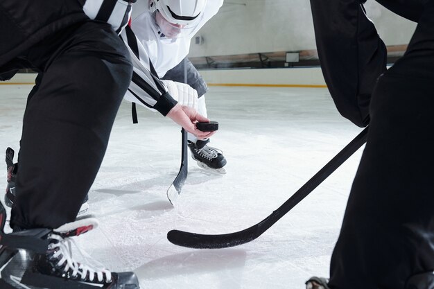 Árbitro de hockey sosteniendo el disco sobre la pista de hielo mientras dos rivales con palos se inclinan hacia adelante y esperan el momento para ser el primero en disparar.