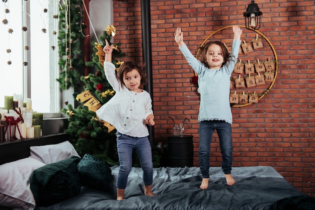 Árbol de Navidad detrás. Niños alegres divirtiéndose y saltando en la cama