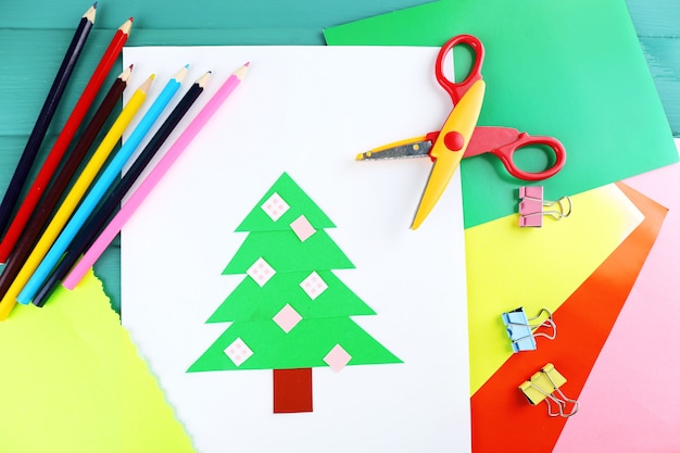 Árbol de Navidad de papel creativo en la hoja de papel blanco, tijeras y crayones de colores en el fondo de la tabla de colores