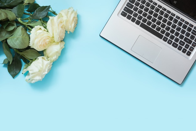 Área de trabalho feminina com laptop e flores rosas brancas na mesa azul.