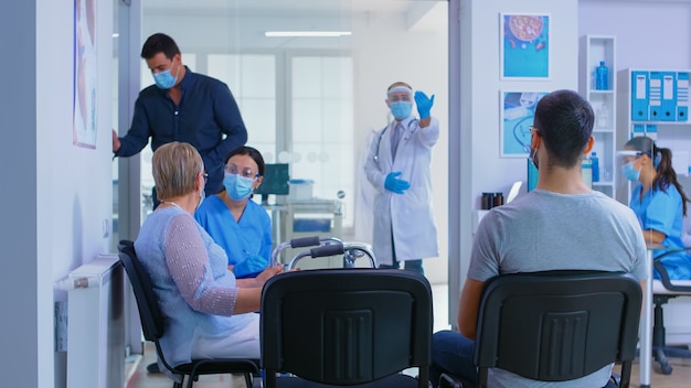 Área de recepción del hospital privado durante la pandemia mundial. Médico con visera contra coronavirus invitando al hombre a su oficina para ser examinado. Mujer mayor discapacitada con andador