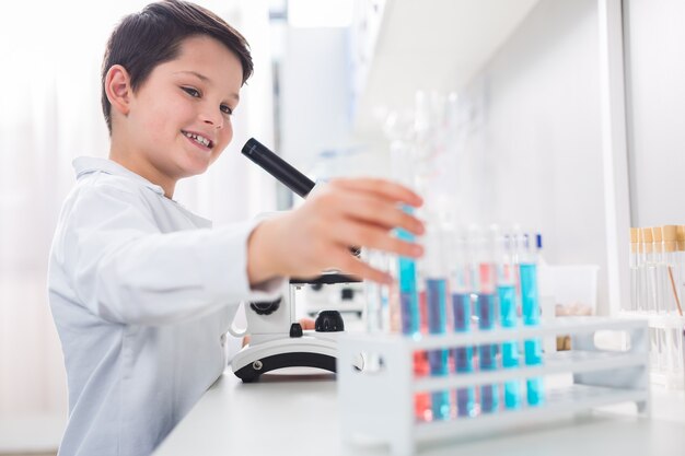 Reactivo apropiado. Chico alegre alegre alegre tomando frasco mientras sonríe y vistiendo bata de laboratorio