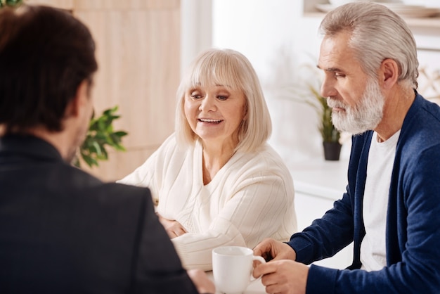 Realización de una compra inmobiliaria. Optimista feliz pareja de ancianos sentados en casa y conversando con el asesor mientras expresan interés