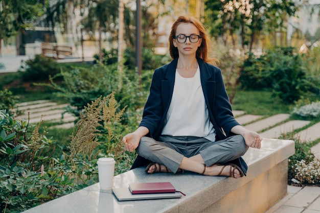 Realxed rothaarige Frau macht Yoga draußen im Park sitzt im Lotussitz meditiert im Freien