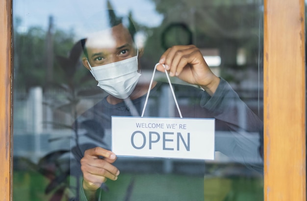 Reapertura de la cafetería después del cierre de covid19 feliz dueño de café afroamericano con máscara médica protectora que abre el letrero en la puerta de vidrio en la cafetería volver al concepto normal