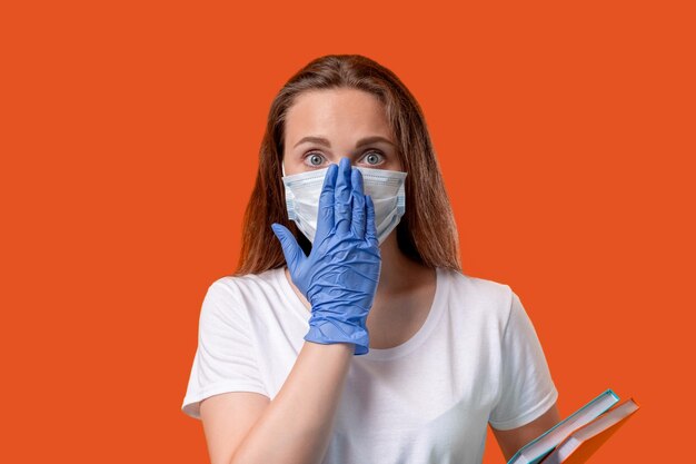 Reapertura de la escuela Educación en cuarentena Estudiante asustada cubriendo la cara con máscara con la mano en guantes protectores aislados en el espacio de copia rojo naranja Pandemia de coronavirus Medidas de higiene Estudio seguro