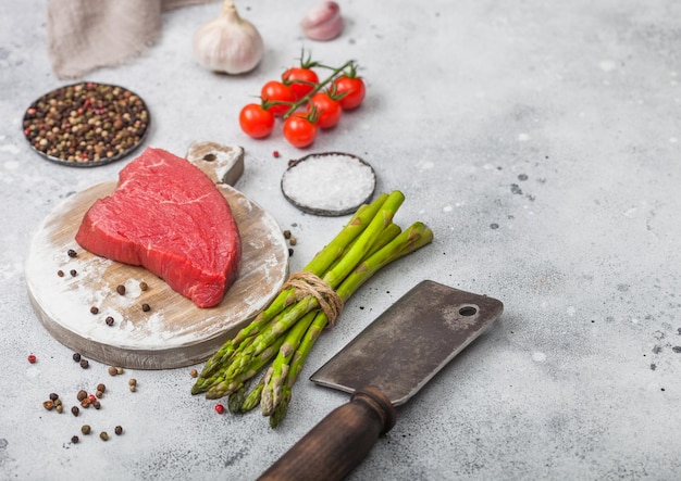 Rebanada de bistec de sirlion de carne cruda en una tabla de cortar redonda con puntas de tomate, ajo y espárragos y hacha de carne en el fondo de la mesa de la cocina clara