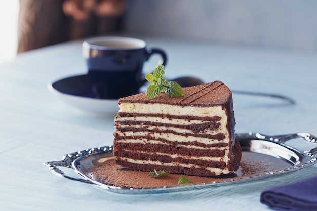 Rebanada de delicioso pastel de chocolate en placa de plata con menta fresca sobre fondo de mesa de madera azul claro.