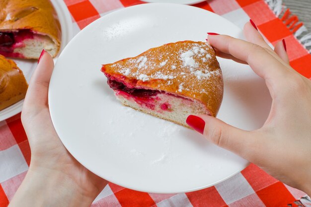 Rebanada de delicioso trozo de tarta de cerezas con azúcar podado en manos de mujer sobre un mantel a cuadros