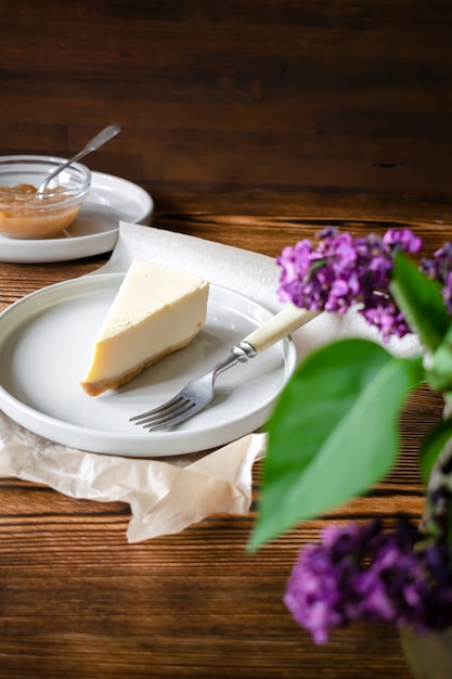 Rebanada de Nueva York clásica del cheesecake del postre dulce delicioso en fondo rústico de madera. Rebanada de pastel sabroso en plato de cerámica servido con tenedor de postre y mermelada de manzana y flores lilas.