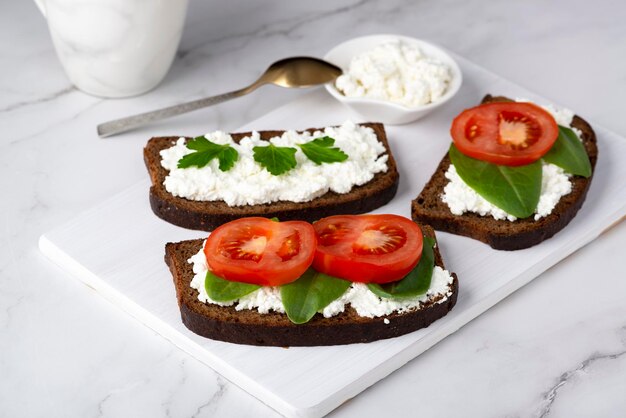 Rebanada de pan de centeno con requesón y tomates en una tabla de cortar de madera sobre un fondo blanco.