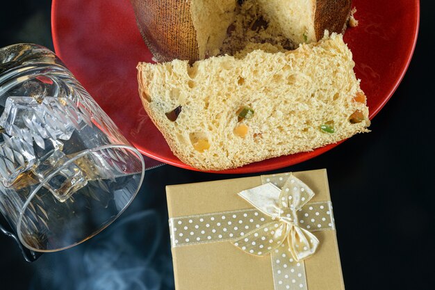 Foto rebanada de panettone junto a un vaso de whisky volcado bajo humo de cirrago y una caja de regalo.