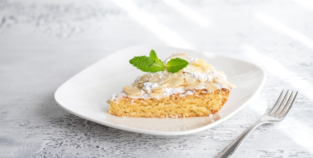 Rebanada de pastel de almendra casera decorada con menta fresca en un plato blanco con luz solar.