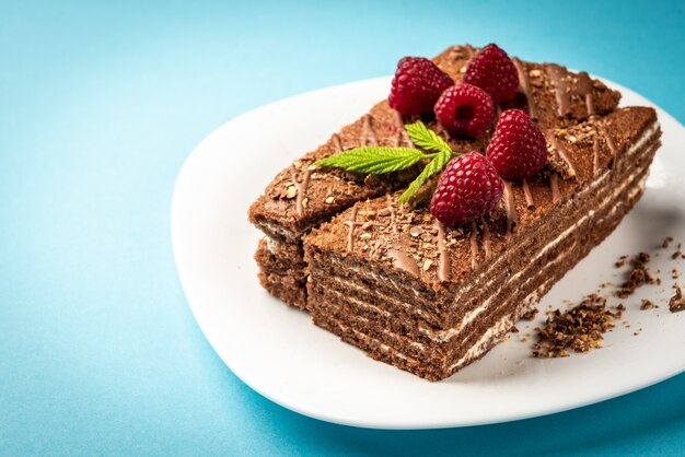 Rebanada de pastel de chocolate con relleno de leche y frambuesa en plato blanco sobre superficie azul.