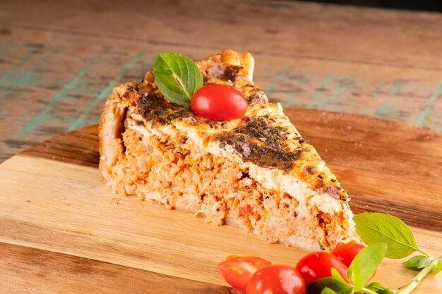 Rebanada de pastel de pollo salado con masa de orégano y tomates cherry sobre una tabla redonda de madera sobre una mesa rústica cerrada en el relleno EN EL LADO