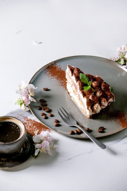 Rebanada de postre italiano tradicional tiramisú sin gluten casero espolvoreado con cacao en polvo decorado con manzano en flor, café, hojas de menta y granos de café sobre la superficie de mármol blanco.