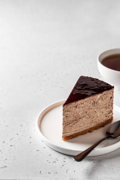 Rebanada de tarta de queso de chocolate glaseada con taza de té sobre fondo blanco texturizado con espacio de texto a la izquierda