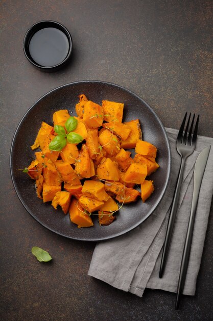 Rebanadas de calabaza frita con hierbas y pimienta, aceite de oliva en una placa de cerámica negra sobre hormigón oscuro o piedra