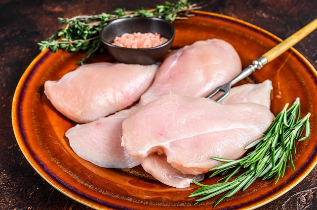 Rebanadas crudas de filetes de filete de pechuga de pollo en un plato rústico con hierbas. Fondo oscuro. Vista superior.