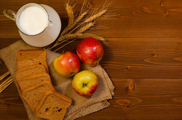Rebanadas de pan de centeno, una jarra de leche, manzanas y mazorcas de maíz sobre tela de saco, mesa de madera