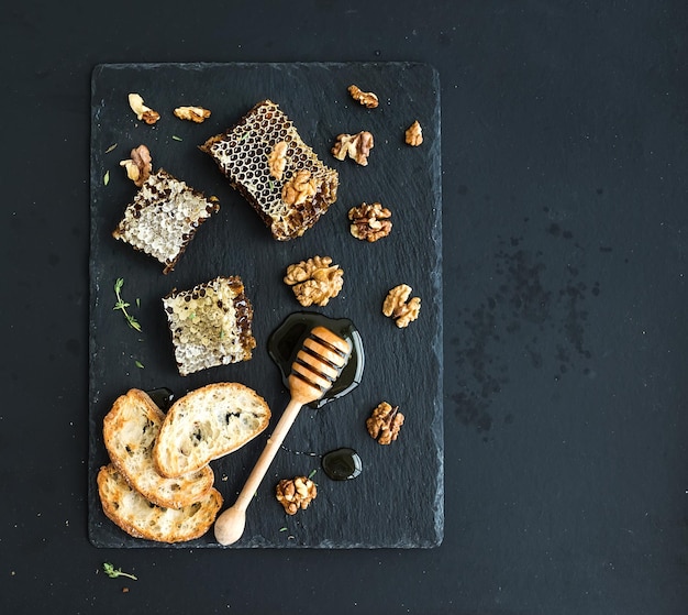 Rebanadas de pan de nueces de panal y cucharón de miel en bandeja de pizarra negra sobre fondo oscuro grunge vista superior