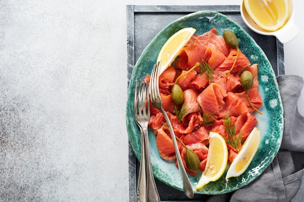 Rebanadas de salmón ligeramente salado con alcaparras, limón y eneldo en un plato de cerámica sobre una mesa de hormigón gris. Vista superior.