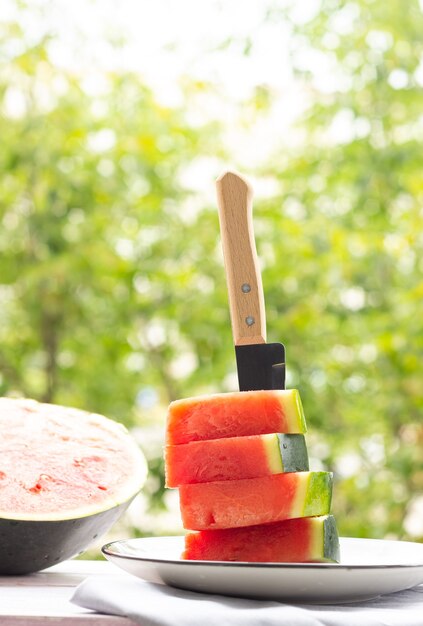 Rebanadas de sandía en un plato con un cuchillo, media sandía en el fondo, al aire libre. Comida sana.