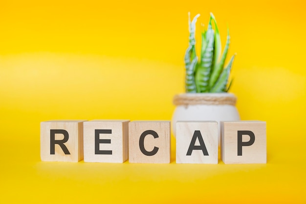 RECAP-Nachricht mit Holzklötzen auf gelbem Hintergrund, grüne Pflanze in einer Blumenvase im Hintergrund