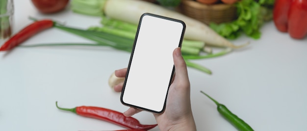 Receita de aparência feminina no smartphone de tela em branco enquanto prepara o ingrediente para cozinhar na mesa da cozinha com legumes frescos