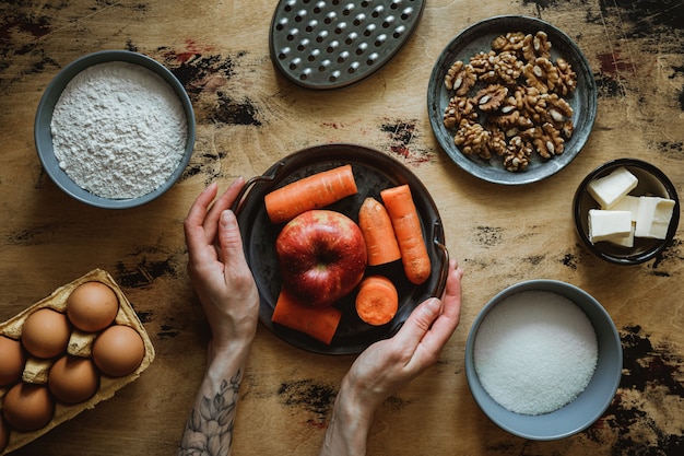 Receita de bolo de pão de noz de cenoura maçã. Ingredientes na mesa de madeira. Farinha, açúcar, manteiga, ovos. Ralador. Mãos na moldura.